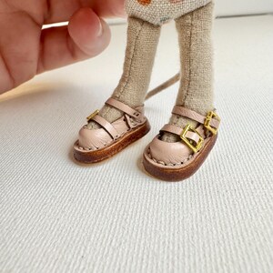 May include: Close-up of miniature doll shoes. The shoes are light pink with two straps and gold buckles. The soles are brown. The doll's legs are tan and the shorts are beige with colorful polka dots.