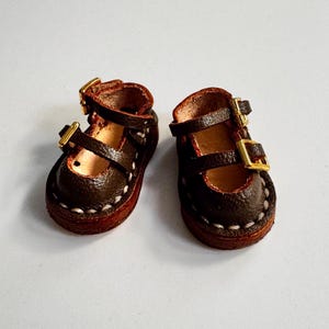 May include: A pair of miniature brown leather Mary Jane shoes with gold-colored buckles. The shoes have a rounded toe and are stitched with white thread around the sole. The shoes are designed for a doll or other small figure.