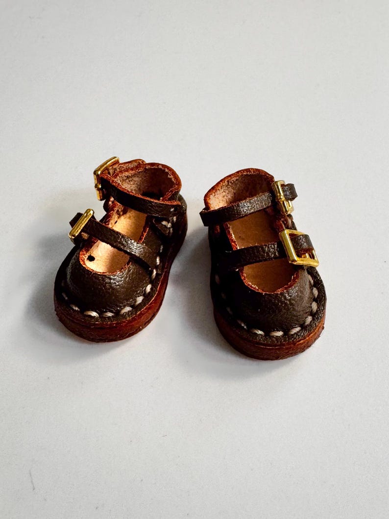 May include: A pair of miniature brown leather Mary Jane shoes with gold-colored buckles. The shoes have a rounded toe and are stitched with white thread around the sole. The shoes are designed for a doll or other small figure.