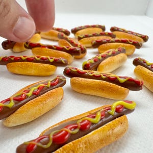 May include: Miniature hot dogs with brown sausages, yellow mustard, and red ketchup on golden-brown buns. A hand is reaching for one of the tiny food items. The food items are arranged on a white surface.