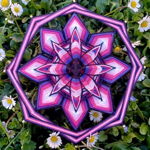 Könnte beinhalten: Ein farbenfrohes Mandala mit rosa, lila, weißem und schwarzem Garn, das zu einem geometrischen Muster gewebt ist. Das Mandala ist von grünem Laub und weißen Blumen umgeben.