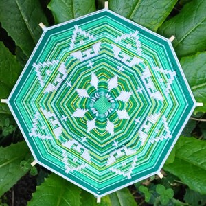 Könnte beinhalten: Ein achteckiger Traumfänger mit einem geometrischen Muster in Grün und Weiß. Der Traumfänger ist aus Garn gefertigt und hat Holzstäbe.