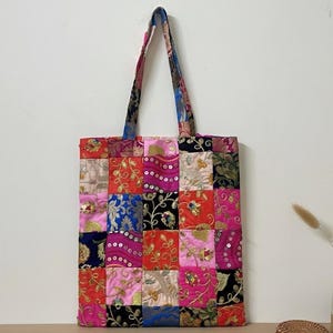 May include: A patchwork tote bag with a colorful design. The bag features a variety of fabrics in squares of pink, red, blue, and gold. The handles are a patterned fabric. The bag is sitting on a wooden surface.
