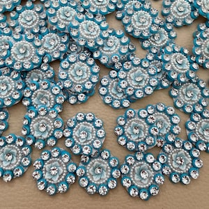 May include: A collection of circular embellishments featuring a central light blue flower surrounded by a ring of clear, faceted rhinestones set in a bright turquoise border. Each piece has a textured white base and a small clear crystal centre.