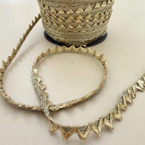 Op de afbeelding: Een spoel met metallic goudkleurige bies met een herhalend driehoekig zaagtandpatroon. Het decoratieve lint is strak om een ronde kern gewikkeld, waarbij een lengte van de gestructureerde, glinsterende gouden stof naar buiten uitsteekt.