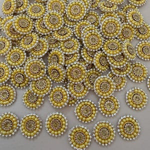 Puede incluir: Una colección de adornos decorativos amarillos y dorados. Cada pieza circular presenta un diseño central rodeado por un anillo de pequeñas cuentas perladas y un anillo exterior de brillantes pedrería transparente. Los adornos están dispersos sobre una superficie neutra.
