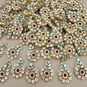 May include: An assortment of gold-coloured, decorative embellishments with red and clear crystal detailing. The designs include floral and leaf motifs, with a central red stone encircled by clear crystals. Some are teardrop-shaped.