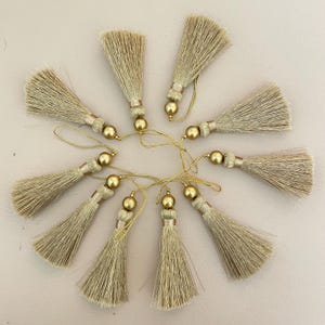 Könnte beinhalten: Zehn beige Quasten mit goldenen Details, kreisförmig angeordnet. Jede Quaste hat eine goldene Perle und eine Schlaufe. Geeignet für verschiedene Bastelprojekte oder Verzierungen.