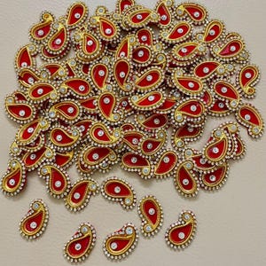 Op de afbeelding: Een verzameling rode en gouden paisley-vormige versieringen. Elk stuk heeft een rood midden, een gouden rand en is bezet met kleine, heldere strass-steentjes. De versieringen zijn verspreid over een neutraal oppervlak.