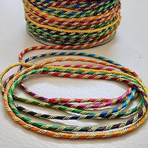 Puede incluir: Un carrete de cordón trenzado multicolor con hilo metálico dorado. Varios hilos del cordón están dispuestos en un patrón circular. El cordón presenta una variedad de colores, incluyendo rojo, azul, verde, amarillo y naranja.