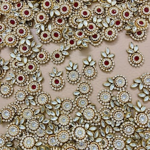 Könnte beinhalten: Eine Auswahl an goldfarbenen Schmuckstücken, darunter Ohrringe und dekorative Elemente. Jedes Stück hat einen zentralen roten Edelstein, der von klaren Kristallen und goldenen Details umgeben ist. Die Designs beinhalten florale und blattartige Muster.
