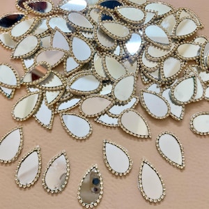 Puede incluir: Una colección de adornos de espejo en forma de lágrima con un borde dorado y un ribete de pedrería. Los espejos están dispersos sobre una superficie rosa claro, reflejando la luz. Ideales para proyectos de manualidades.