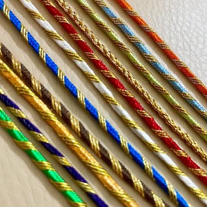 Puede incluir: Primer plano de cordones trenzados coloridos con detalles metálicos dorados. Los cordones presentan varias combinaciones de colores, incluyendo rojo y blanco, azul y marrón, y verde y dorado. Los cordones están dispuestos en diagonal, mostrando su intrincado diseño.