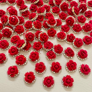 Puede incluir: Una colección de pequeños adornos de rosas de terciopelo rojo con un borde de pedrería brillante. Estos acentos florales decorativos están dispersos sobre una superficie clara, mostrando sus intrincados detalles y su color vibrante.