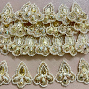 May include: Close-up of multiple cream-colored, beaded appliques. Each applique features a central teardrop-shaped gem, surrounded by smaller gold and iridescent beads. The edges are outlined with gold thread, creating a detailed, ornate design.