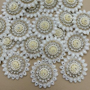 May include: A collection of circular, flower-shaped patches featuring a central cream-coloured floral element surrounded by concentric rings of small white pearls and clear rhinestones, finished with a scalloped white embroidered border.