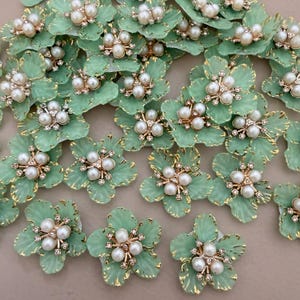 May include: A collection of mint green floral embellishments with gold trim. Each flower features a cluster of white pearls and small clear rhinestones at the center. The petals have a delicate, veined texture.
