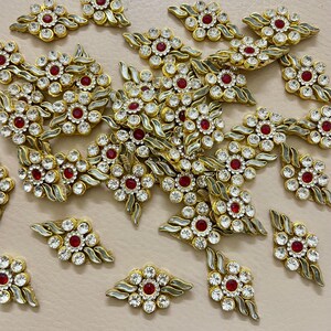 May include: Close-up of multiple gold-toned floral embellishments. Each piece features a central red gemstone surrounded by clear crystals. The embellishments have a leaf-like design and are arranged on a light beige surface.