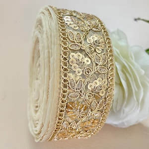 Peut inclure: Un rouleau de dentelle ivoire avec des broderies florales dorées et des sequins. La garniture présente un motif détaillé de feuilles et de fleurs, idéal pour embellir des vêtements ou des créations artisanales.