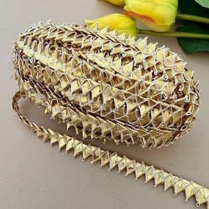May include: A roll of gold metallic trim featuring a geometric design and white stitching. The trim has a scalloped edge, ideal for craft and sewing projects. Yellow flowers are visible in the background.