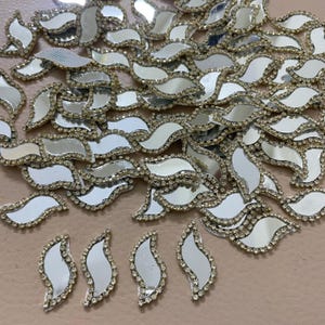 May include: A collection of silver, leaf-shaped embellishments with a mirrored surface and a border of small, clear rhinestones. The decorative pieces are scattered across a neutral surface, showcasing their reflective quality.