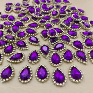 May include: A collection of teardrop-shaped purple rhinestones with a gold-colored border and small clear rhinestones. These decorative embellishments are ideal for adding sparkle to crafts, clothing, or accessories.