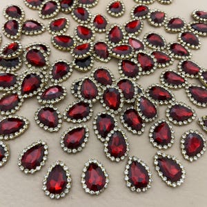 Op de afbeelding: Een verzameling rode, druppelvormige strass-steentjes met een goudkleurige rand en een omranding van kleine, heldere strass-steentjes. Deze decoratieve versieringen zijn verspreid over een neutrale ondergrond.