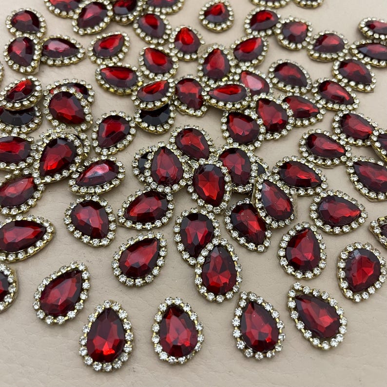 Op de afbeelding: Een verzameling rode, druppelvormige strass-steentjes met een goudkleurige rand en een omranding van kleine, heldere strass-steentjes. Deze decoratieve versieringen zijn verspreid over een neutrale ondergrond.