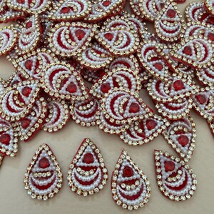 May include: A collection of ornate, teardrop-shaped embellishments. Each piece features a red center stone, surrounded by white faux pearls, gold-colored trim, and clear rhinestones. The pieces are scattered across a neutral surface.
