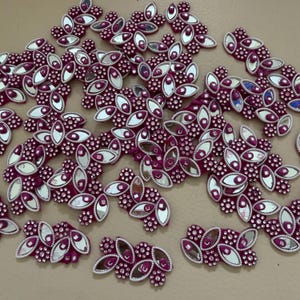 Pode incluir: Uma coleção de enfeites decorativos com um design floral. Cada peça apresenta uma base magenta escura com detalhes ovais espelhados e pequenas pedras de strass transparentes. Os enfeites estão espalhados sobre uma superfície neutra.