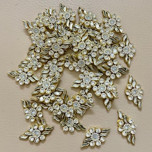 May include: A selection of gold-coloured floral embellishments, each with a central cluster of clear rhinestones. The embellishments feature a leaf design and are arranged on a neutral surface, suitable for crafting.