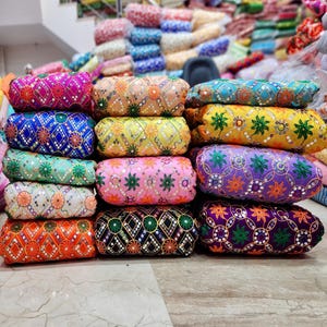 May include: A display of embroidered fabric rolls in a variety of colours. The rolls showcase detailed patterns with shades of pink, blue, orange, and green. The designs include floral and geometric motifs, with reflective embellishments.