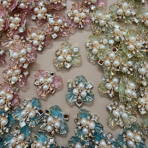 May include: A collection of flower-shaped embellishments in shades of pink, blue, and green. Each piece has a cluster of faux pearls at the centre, surrounded by clear and gold-toned details. These decorative elements are suitable for crafting and jewellery making.