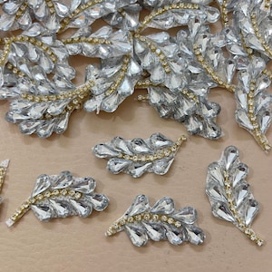 Może przedstawiać: Zbliżenie kilku dekoracyjnych ozdób w kształcie liści. Każdy liść ma przezroczyste, fasetowane kamienie przypominające kryształ i złote akcenty. Liście ułożone są na jasnobrązowej powierzchni, tworząc efekt błyszczący i elegancki.