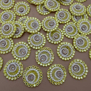 May include: A collection of yellow, white, and gold circular embellishments. Each has a spiral design with a central jewel and is surrounded by beads and small, square-cut gems. The background is a solid, neutral color.