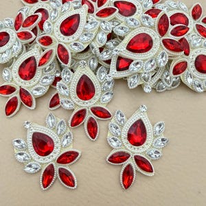 Puede incluir: Una colección de apliques en forma de lágrima con una piedra roja facetada central rodeada de cristales blancos transparentes más pequeños. Cada pieza está dispuesta en un patrón floral con un borde de cuentas blancas para decoración.