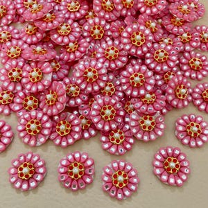 May include: A collection of pink flower-shaped embellishments with gold and pearl accents. Each flower has a central pearl surrounded by red and gold details, with a border of small, reflective squares. These decorative elements are ideal for craft projects.