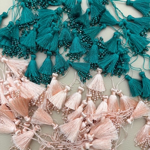 May include: A collection of numerous small tassels in teal and pale pink. Each tassel features a dense cluster of fine threads topped with a matching cord loop and is accented with small, shimmering beads integrated into the lower fringe.