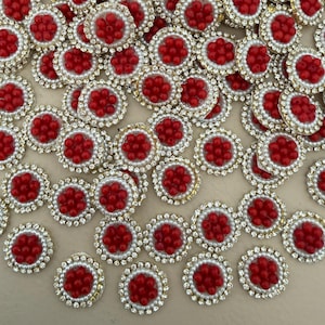 Puede incluir: Una colección de adornos decorativos. Cada adorno circular presenta un grupo central de cuentas rojas, rodeado por un anillo de perlas blancas y un borde de pedrería transparente engastada en metal dorado.