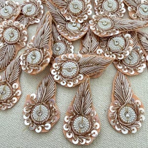 May include: Close-up of several decorative fabric appliques. Each applique features a central beaded design surrounded by sequins and metallic thread. The appliques have a teardrop or leaf shape with a peach-colored fabric backing.