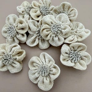 Puede incluir: Una colección de siete flores artesanales hechas de tela de arpillera de color beige claro. Cada flor presenta un centro circular con una textura tejida apretada rodeada por múltiples pétalos redondeados y fruncidos hechos de la misma fibra natural.