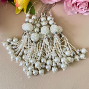 Puede incluir: Un conjunto de borlas de perlas blancas con detalles plateados. Cada borla presenta una gran esfera con cuentas en la parte superior, con hilos de perlas más pequeñas que caen en cascada. Las borlas están dispuestas sobre una superficie rosa claro, con flores de fondo.