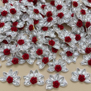 May include: A collection of decorative floral appliques featuring a central red rose surrounded by a fan-shaped arrangement of clear, marquise-cut crystals set on a white fabric backing. Each piece is designed with a symmetrical petal-like layout.