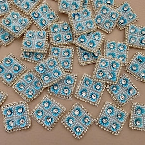 May include: A selection of square embellishments featuring a light blue floral pattern and sparkling rhinestones. Each square has a gold-coloured border. These decorative pieces are suitable for craft projects and are set against a neutral background.