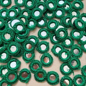 May include: A collection of small, circular embellishments. Each piece features a central mirror surrounded by a ring of bright green thread. The items are scattered, creating a textured, decorative effect. Ideal for craft projects.