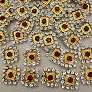 Puede incluir: Una colección de parches decorativos cuadrados con una piedra preciosa circular de color rojo oscuro en el centro, engastada en un marco de metal dorado, rodeada por un borde de pequeños cristales blancos facetados en patrón cuadrado.