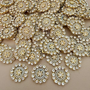 May include: A collection of gold-toned, circular embellishments with intricate detailing. Each piece features a central cluster of clear, sparkling rhinestones surrounded by smaller, pearl-like beads and additional rhinestones. The overall design is ornate and decorative.