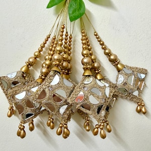 Può includere: Nappe decorative dorate con dettagli a specchio. Ogni nappa presenta un design a forma di stella con piccoli specchi, ricami dorati e perline dorate pendenti. Le nappe sono sospese da fili di perline dorate.