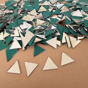 May include: A collection of small, mirrored glass triangles with silver edges and a green backing. The triangles are scattered across a tan surface, creating a mosaic effect. Ideal for craft projects.