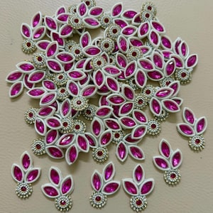 May include: A collection of decorative embellishments featuring a leaf-like design. Each piece has a white base with vibrant pink, oval-shaped gemstones and a central red gemstone. The embellishments are accented with silver-colored beads.